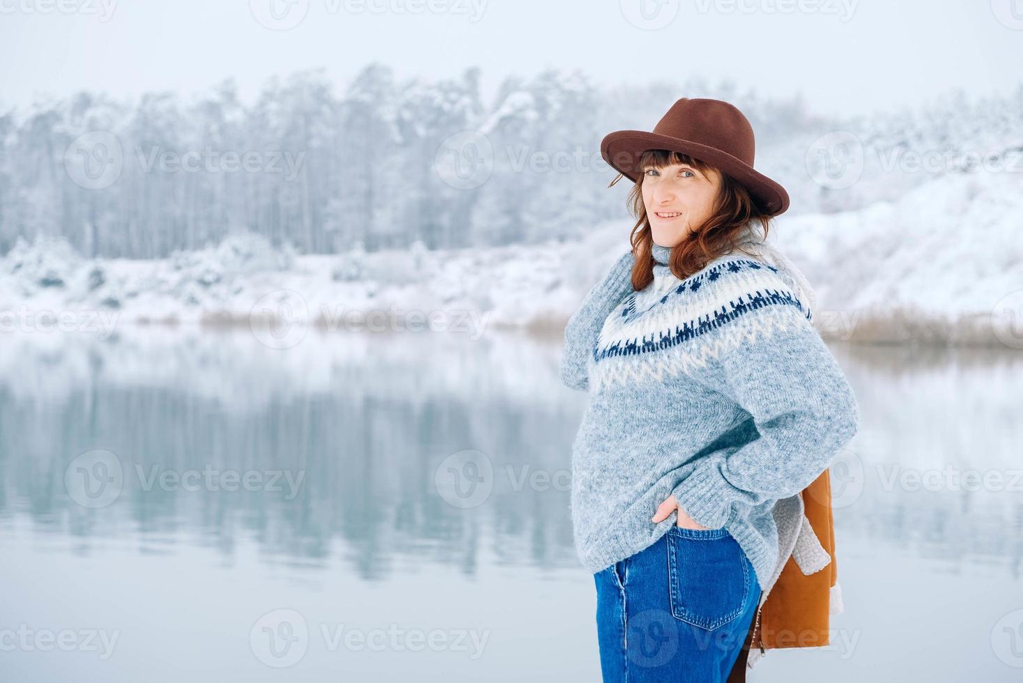 mulher vestindo chapéu e jaqueta quente em pé nas margens de um lago coberto de neve e floresta foto