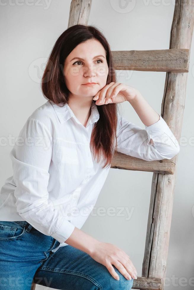 linda mulher vestida com uma camisa branca e jeans azul posando perto de uma escada de madeira em um fundo branco foto