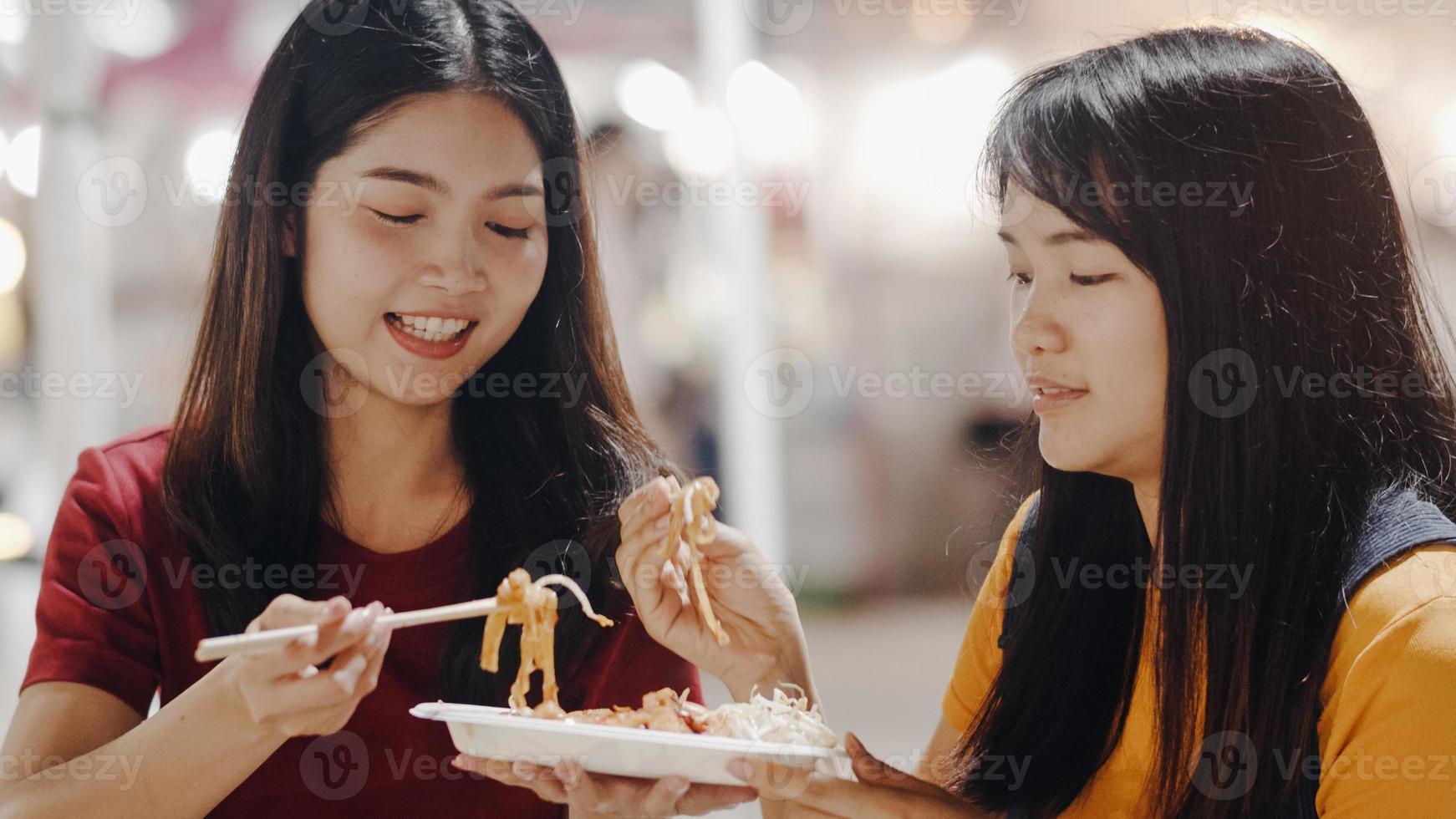 amigos de mulheres jovens asiáticas viajam em bangkok, tailândia, linda fêmea se sentindo feliz andando e comendo pad thai na estrada khao san. as mulheres viajam comem comida de rua no conceito de tailândia. foto