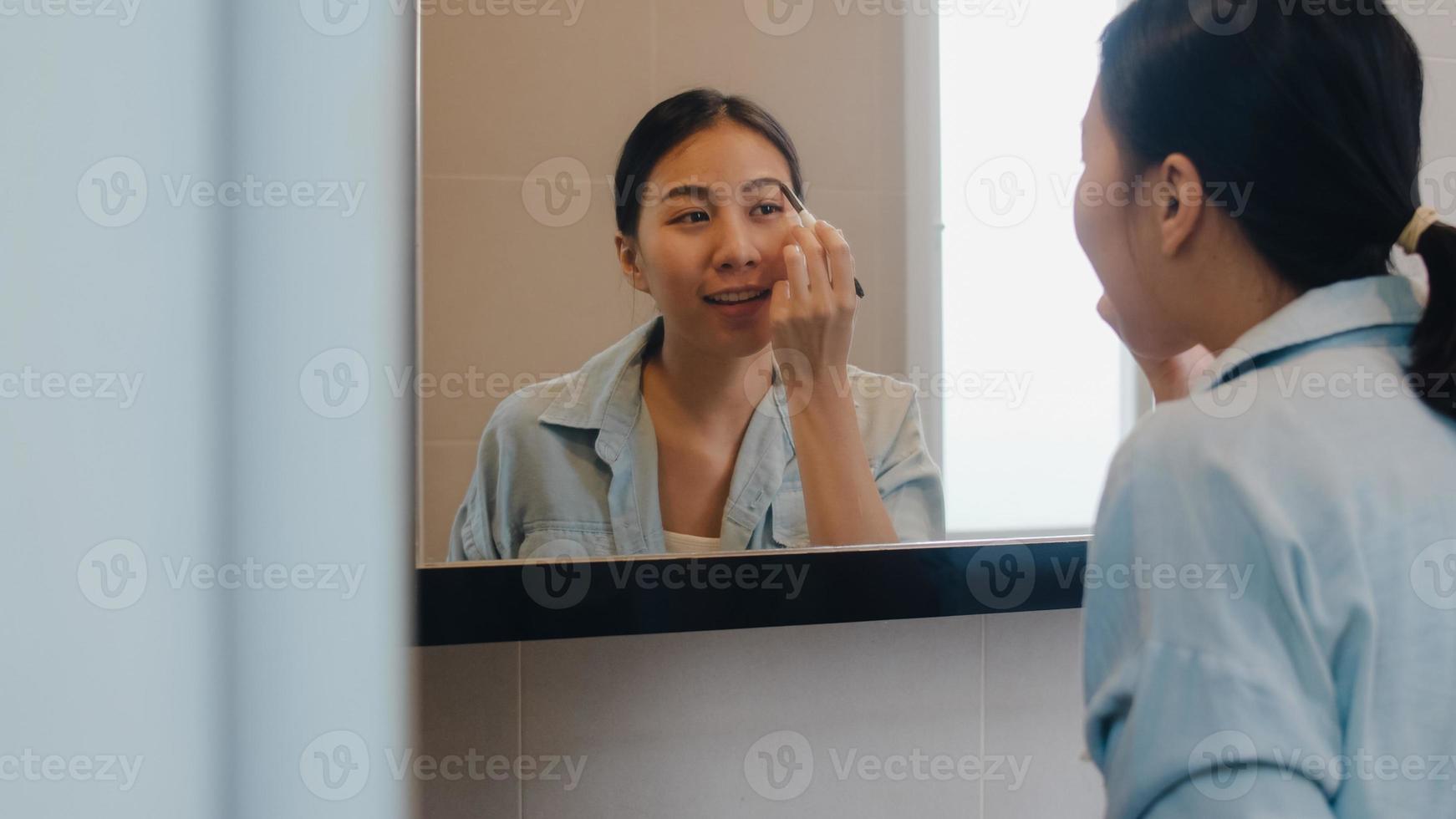 linda mulher asiática usando lápis de sobrancelha maquiagem no espelho frontal, mulher latina feliz usando cosméticos de beleza para melhorar-se pronta para trabalhar no banheiro em casa. mulheres de estilo de vida relaxam em casa. foto