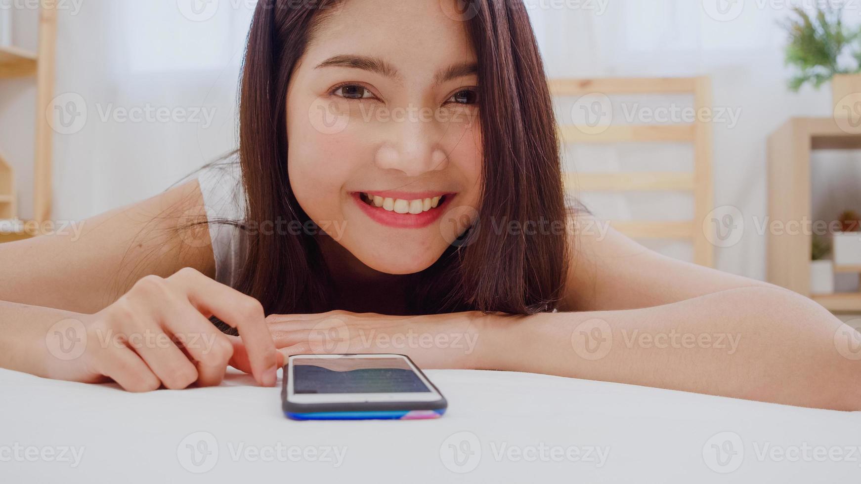 jovem mulher asiática usando smartphone verificando as mídias sociais, sentindo-se feliz sorrindo enquanto estava deitado na cama depois de acordar de manhã, linda garota japonesa atraente sorrindo relaxa no quarto em casa. foto