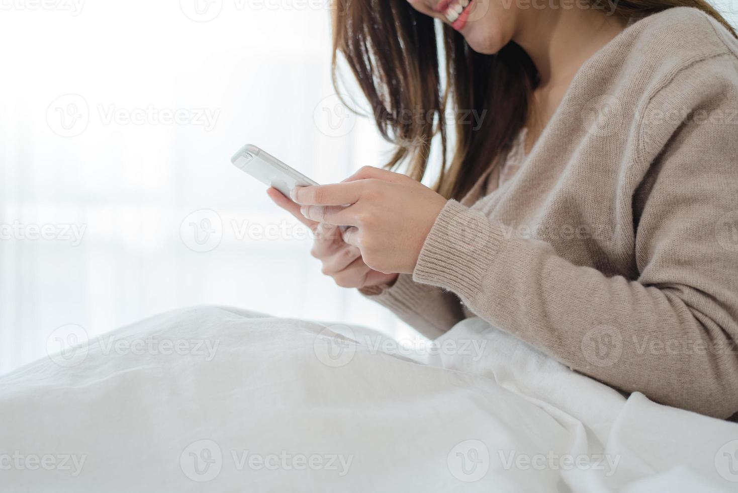 mulheres asiáticas felizes estão usando o telefone inteligente na cama pela manhã. mulher asiática na cama, verificando aplicativos sociais com o smartphone. mulher sorridente navegando na net com o celular em casa. conceito de viciado em celular. foto