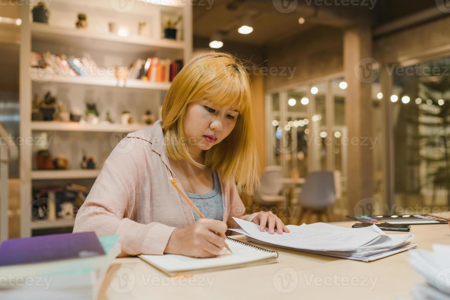 mulheres estudantes asiáticas lendo livros na biblioteca da universidade. jovem estudante de graduação faz lição de casa, lê livro didático, estuda muito para obter conhecimento na mesa de aula na noite de horas extras do campus da faculdade. foto