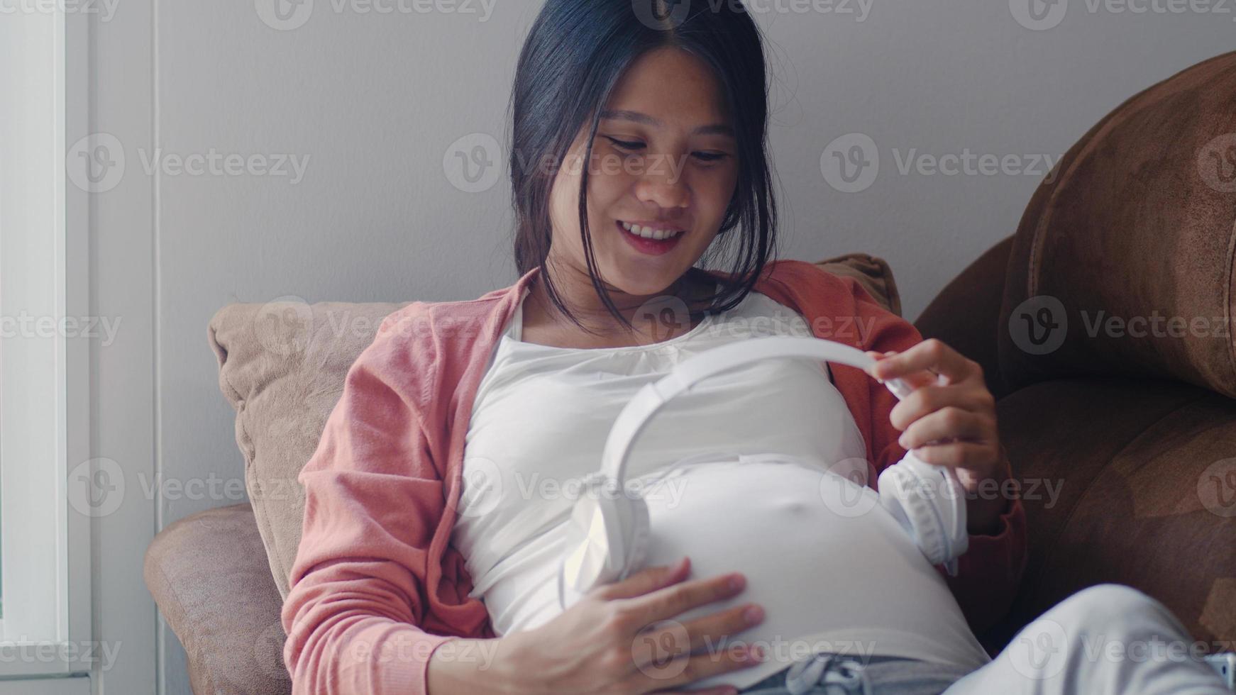 jovem mulher grávida asiática usando telefone e fone de ouvido toca música para bebê na barriga. mãe se sentindo feliz sorrindo positivo e pacífico enquanto cuida da criança deitada no sofá na sala de estar em casa conceito. foto