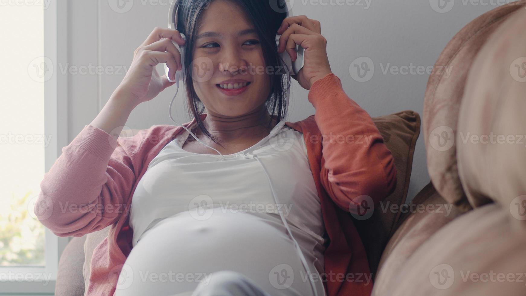 jovem mulher grávida asiática usando telefone e fone de ouvido toca música para bebê na barriga. mãe se sentindo feliz sorrindo positivo e pacífico enquanto cuida da criança deitada no sofá na sala de estar em casa conceito. foto