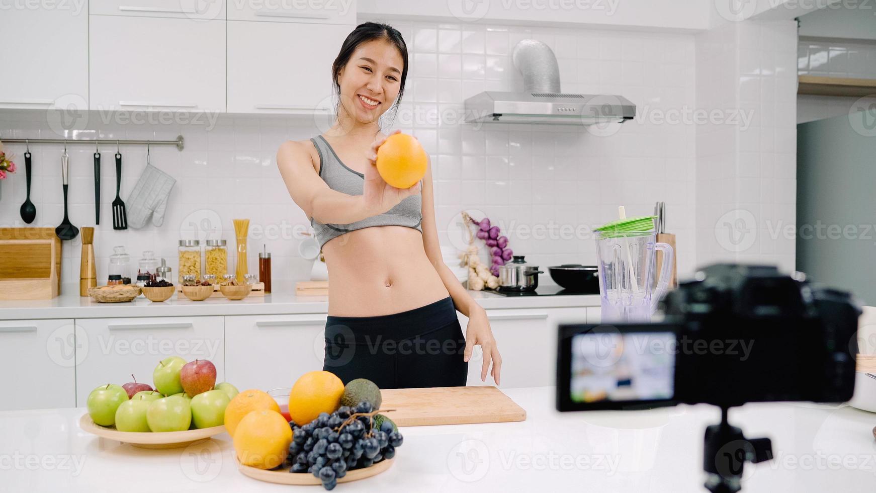 blogueira esportiva mulher asiática usando câmera gravando como fazer um vídeo de suco de laranja para seu assinante, mulher usa frutas orgânicas fazendo suco de laranja sozinha em casa. conceito de comida saudável. foto