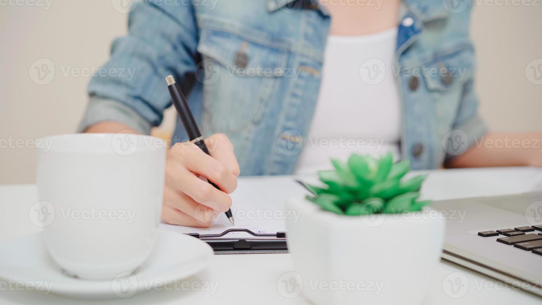 bela jovem sorridente mulher asiática trabalhando laptop na mesa na sala de estar em casa. mulher de negócios da Ásia escrevendo finanças de documento de caderno e calculadora no escritório doméstico. aproveitando o tempo no conceito de casa. foto