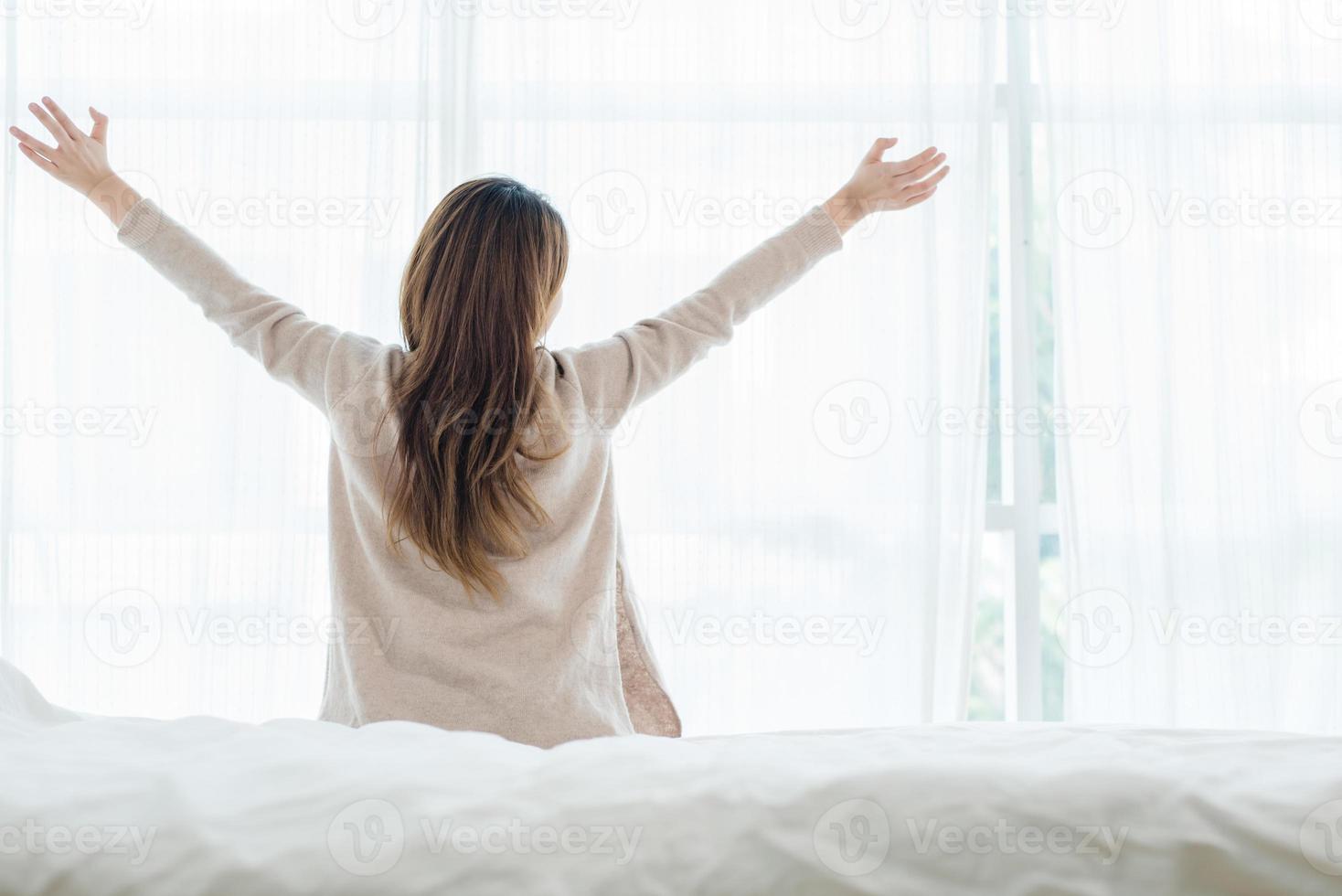 vista traseira da bela jovem asiática feliz acordando de manhã, sentado na cama, esticando-se no quarto aconchegante, olhando pela janela. mulher engraçada depois de acordar. ela está esticando e sorrindo. foto