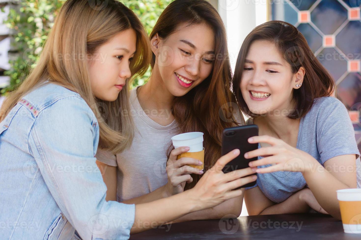 alegres mulheres jovens asiáticas sentadas no café tomando café com amigos e conversando juntos. mulher asiática atraente desfrutando de café enquanto estiver usando o smartphone para falar, ler e enviar mensagens de texto. foto
