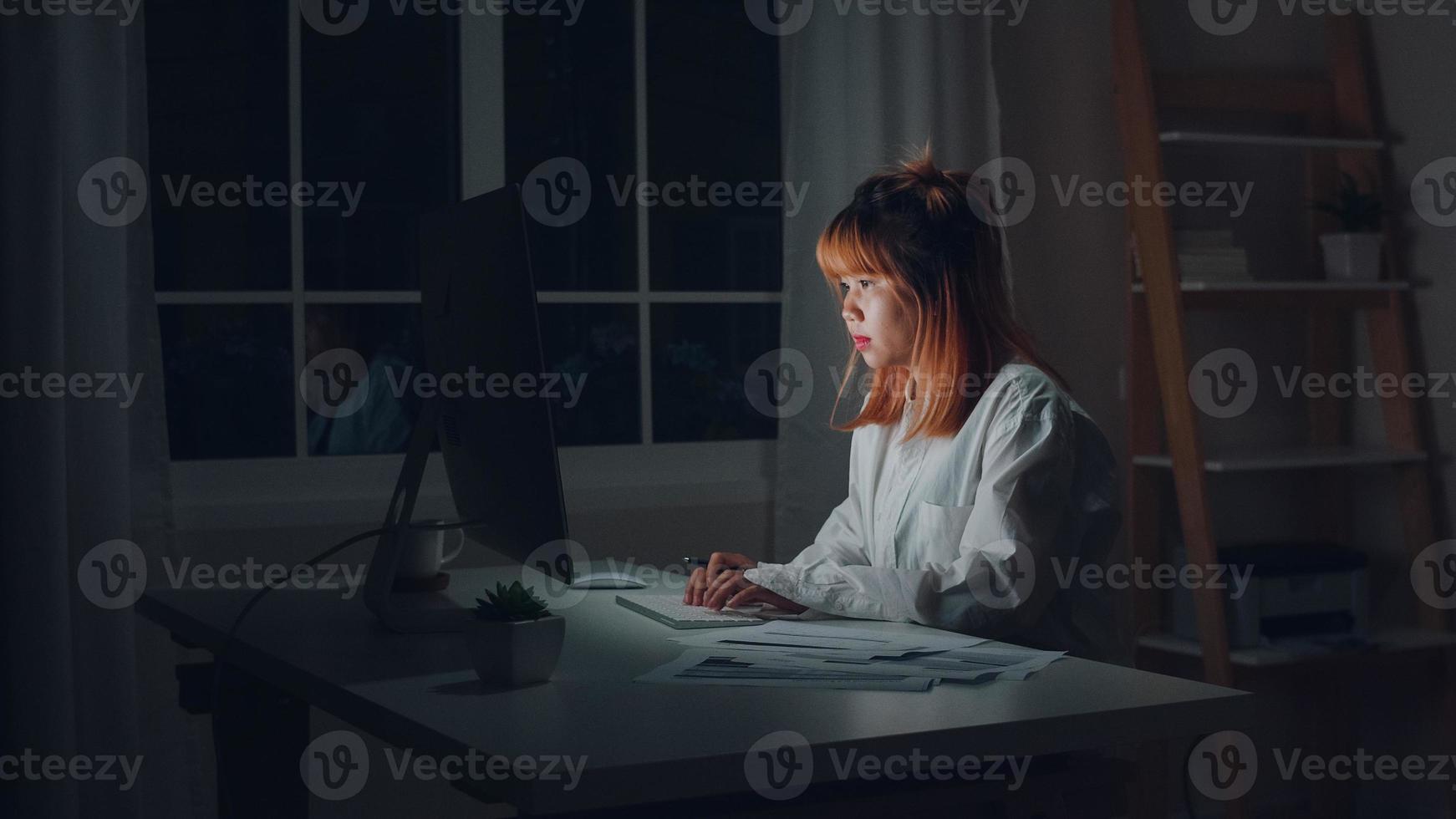 jovem mulher asiática trabalhando até tarde usando o desktop na mesa na sala de estar em casa. mulher de negócios da ásia escrevendo finanças de documento de caderno e calculadora à noite no escritório em casa. conceito feminino sobrecarregado. foto