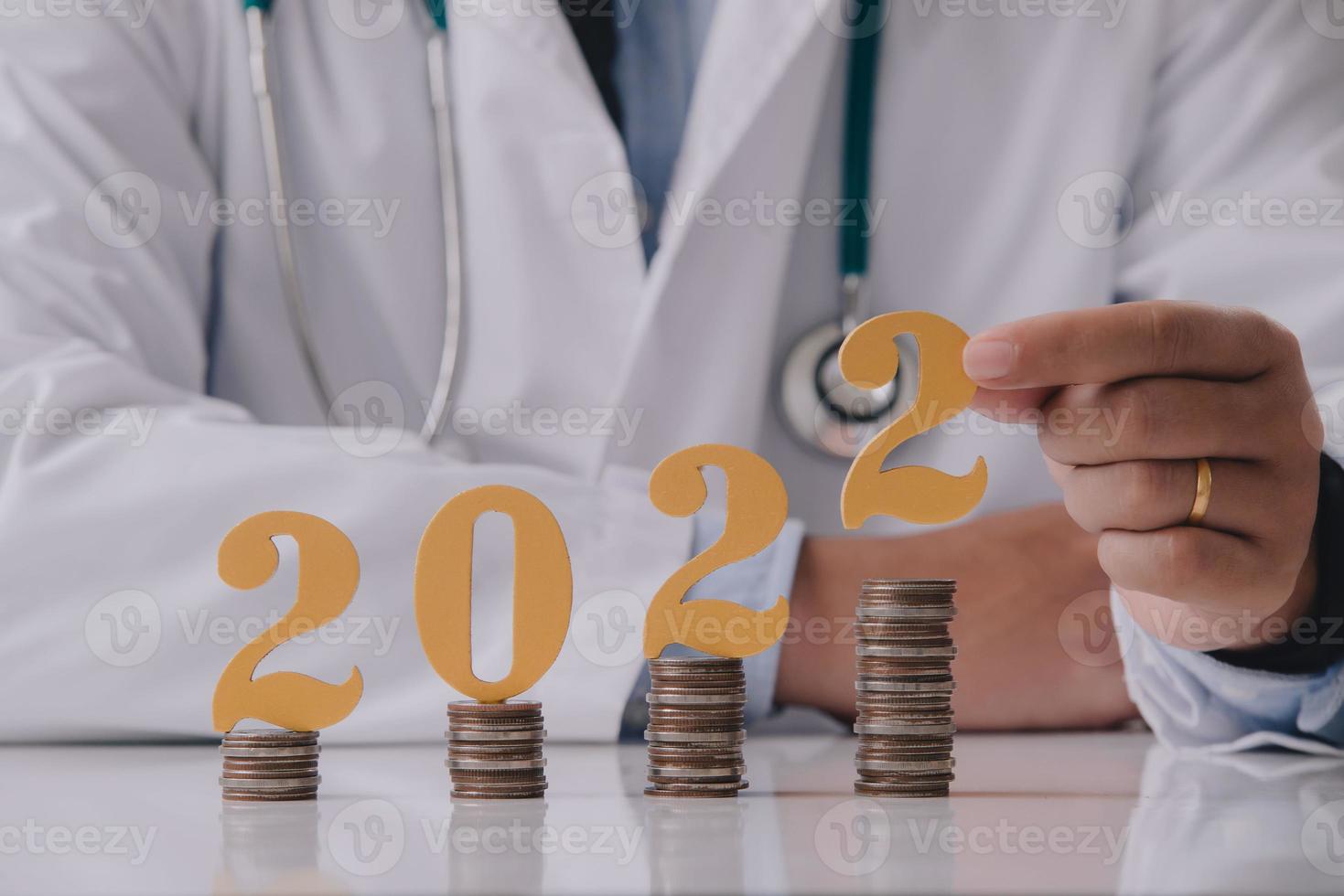 médico de uniforme branco segurando o número 2022 com moeda na mesa do médico. feliz ano novo para cuidados de saúde e conceito médico. foto