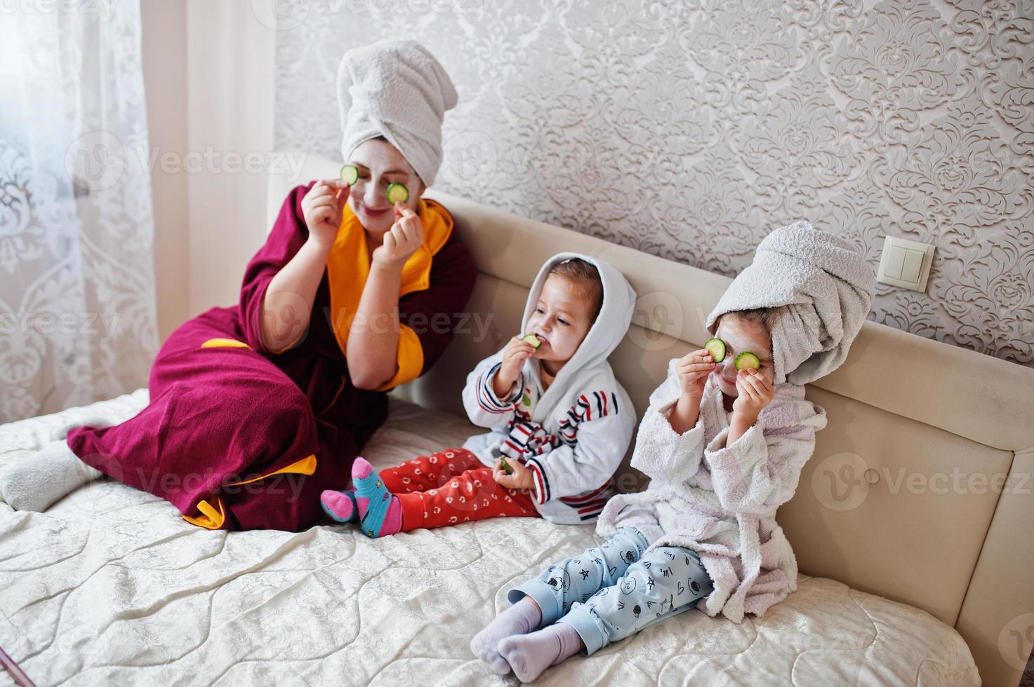 mãe com filhas em um roupão de banho com uma toalha na cabeça cuida da pele em casa, pepinos nos olhos. foto