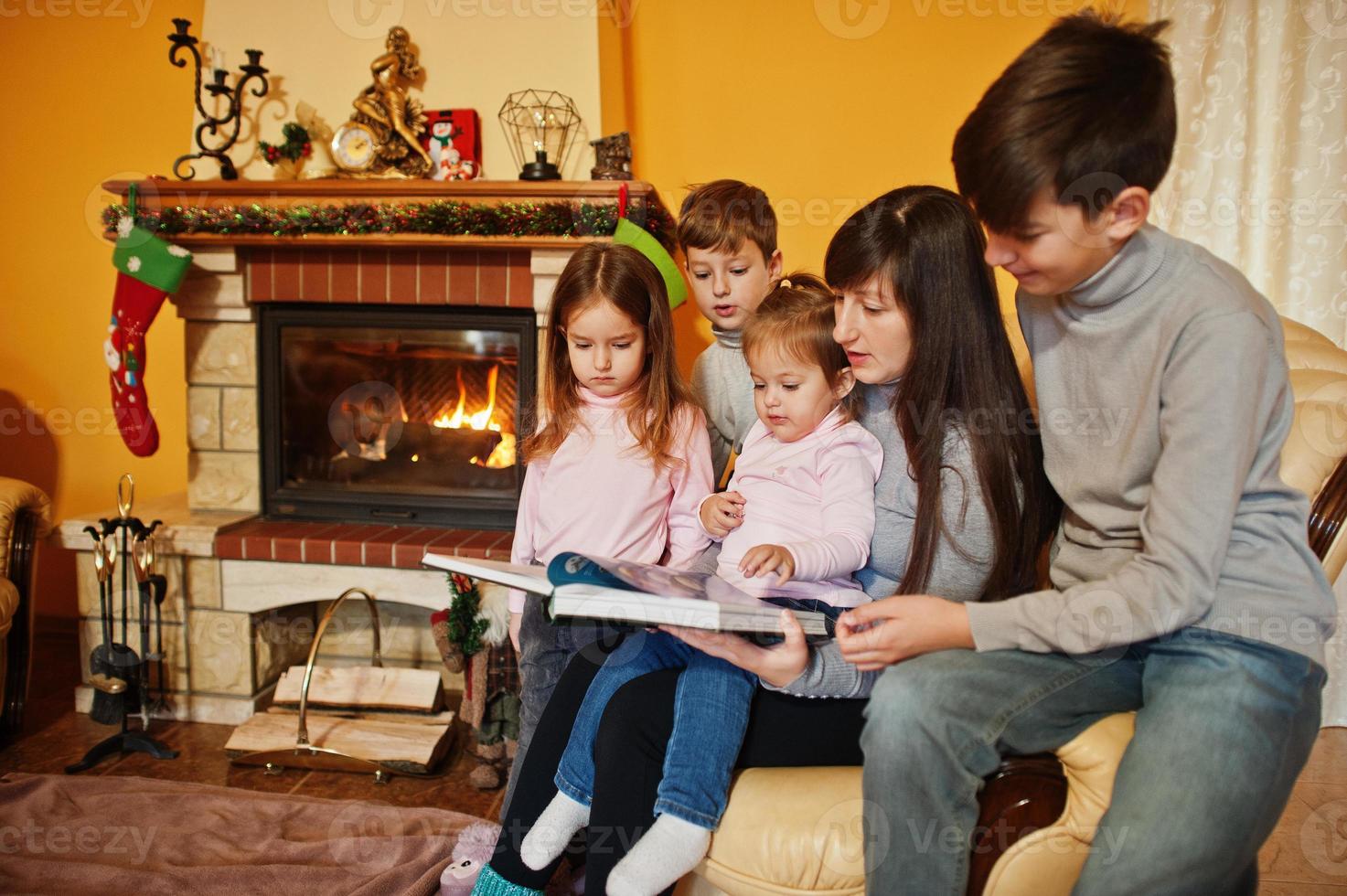 feliz jovem grande família por uma lareira na sala de estar quente em dia de inverno. mãe com quatro filhos em casa leu o livro. foto