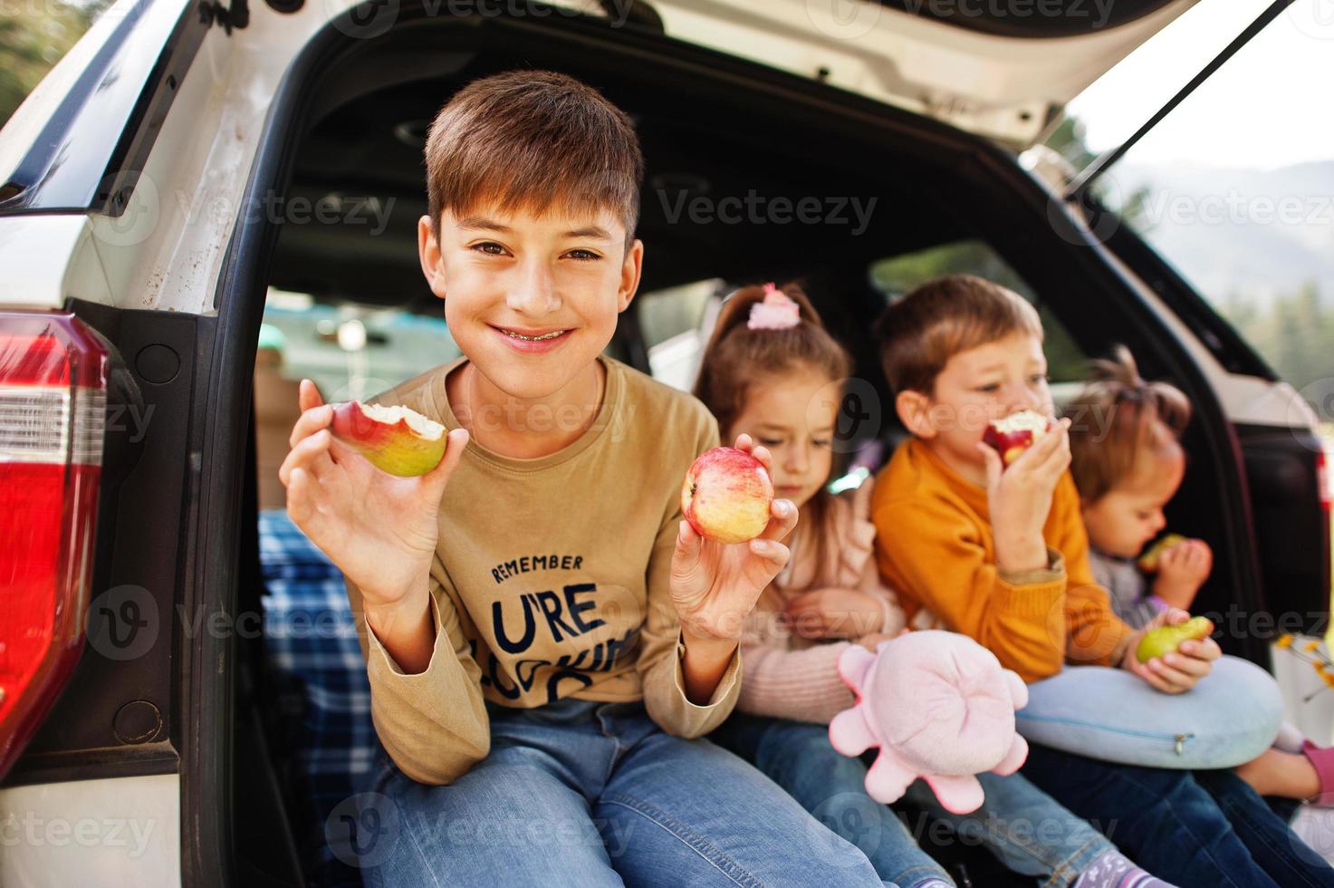 família de quatro filhos come maçãs no interior do veículo. crianças sentadas no porta-malas. viajando de carro nas montanhas, conceito de atmosfera. foto