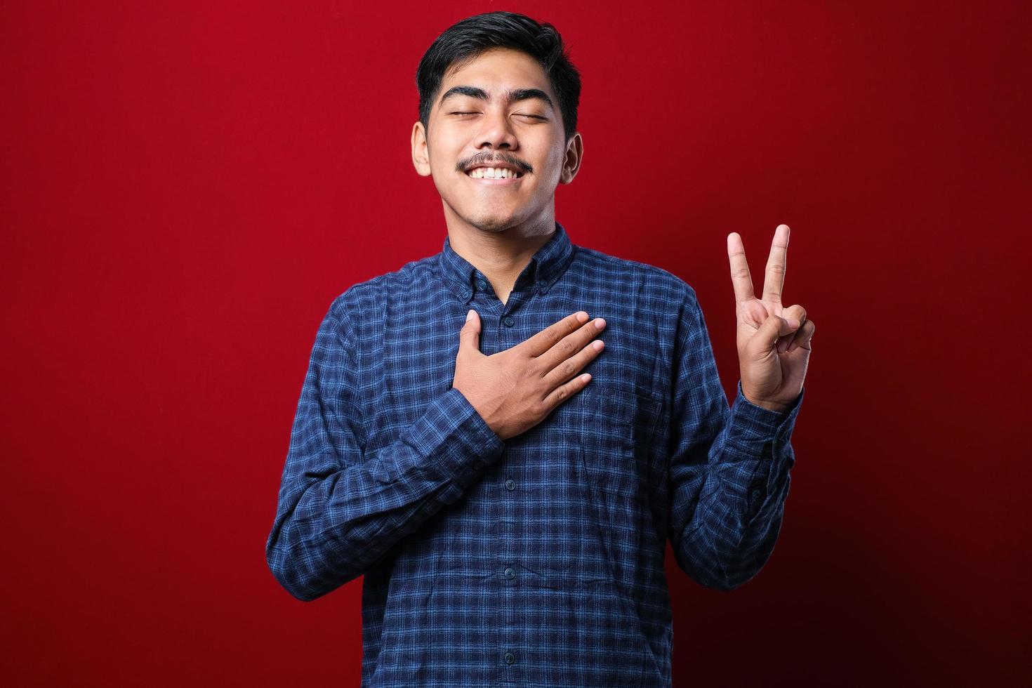 jovem asiático bonito vestindo camisa casual sorrindo xingando com a mão no peito e dedos para cima, fazendo um juramento de promessa de lealdade foto