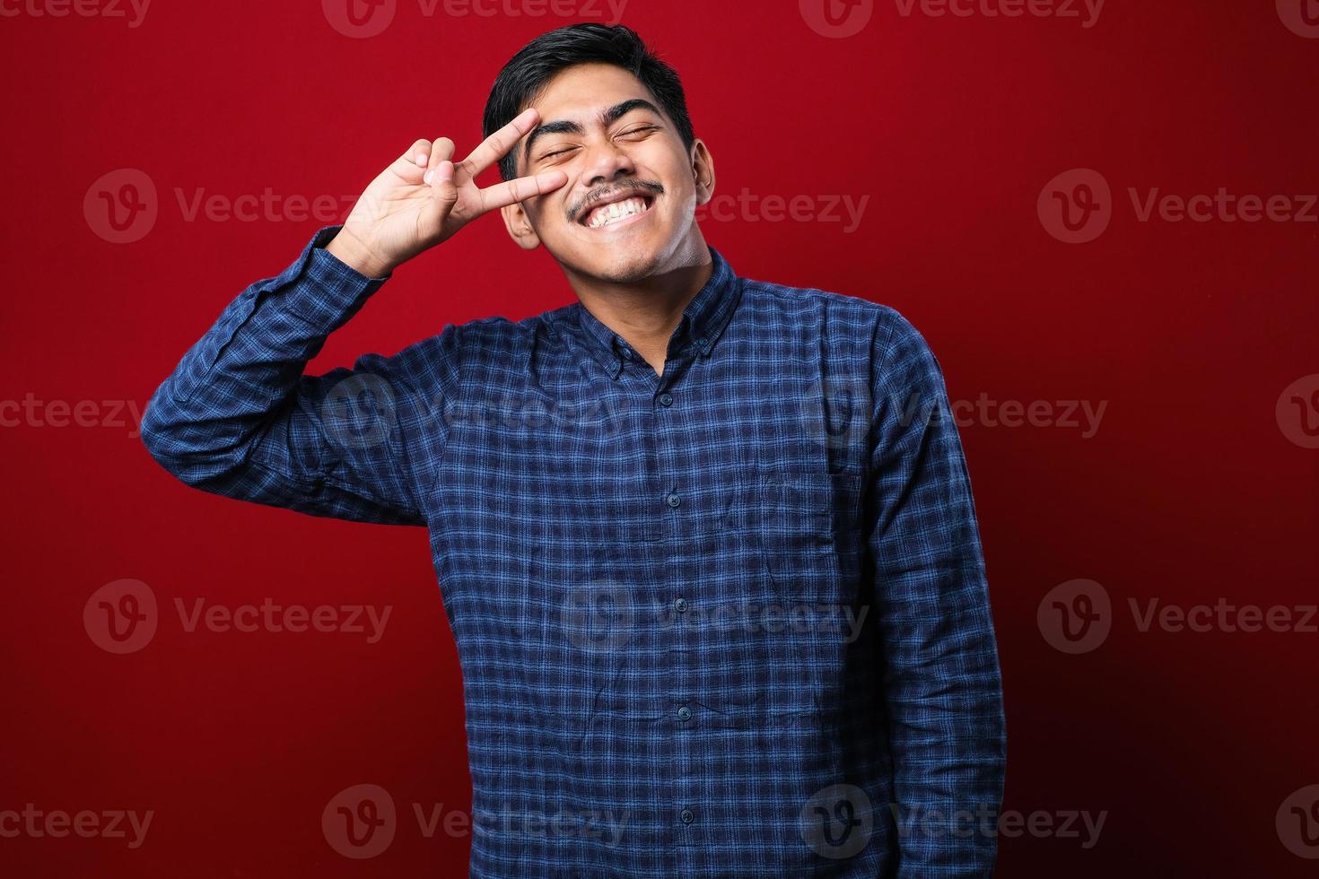 jovem asiático bonito vestindo camisa casual, fazendo o símbolo da paz com os dedos sobre o rosto, sorrindo alegre mostrando vitória foto