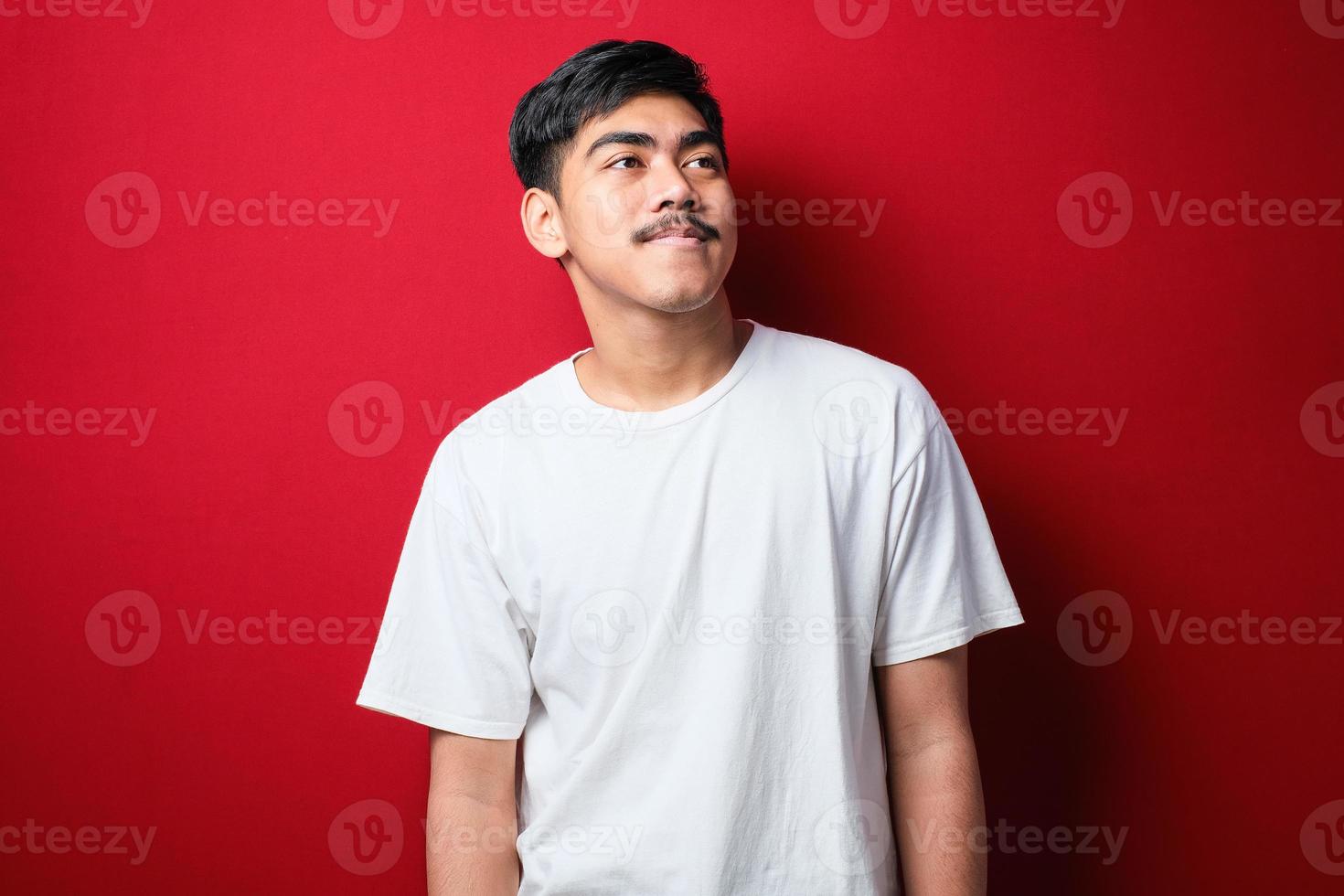 jovem bonito vestindo camiseta branca sobre fundo vermelho isolado, olhando para o lado com um sorriso no rosto, expressão natural. foto