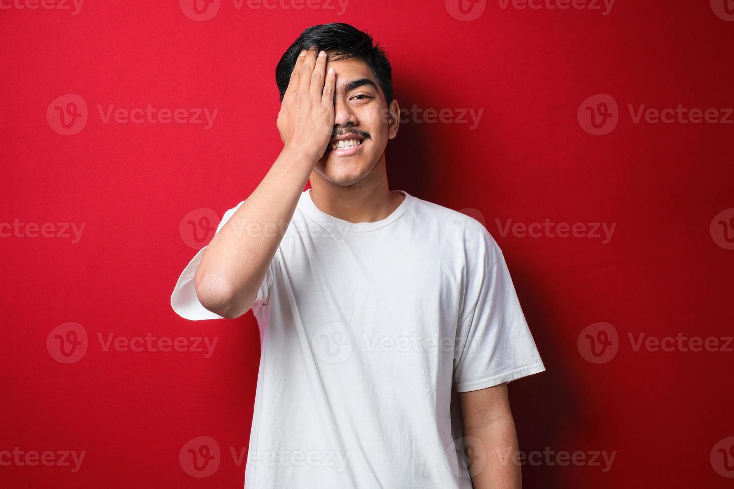 jovem asiático vestindo camiseta branca sorrindo e rindo com a mão no rosto cobrindo os olhos de surpresa. conceito cego foto