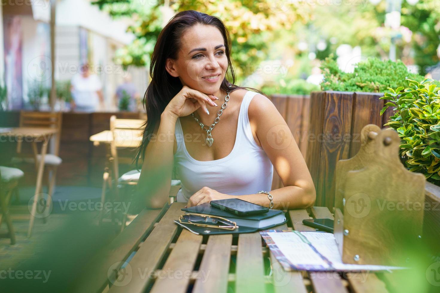 jovem satisfeita em t-shirt branca senta-se à mesa de um café de rua ao ar livre. foto