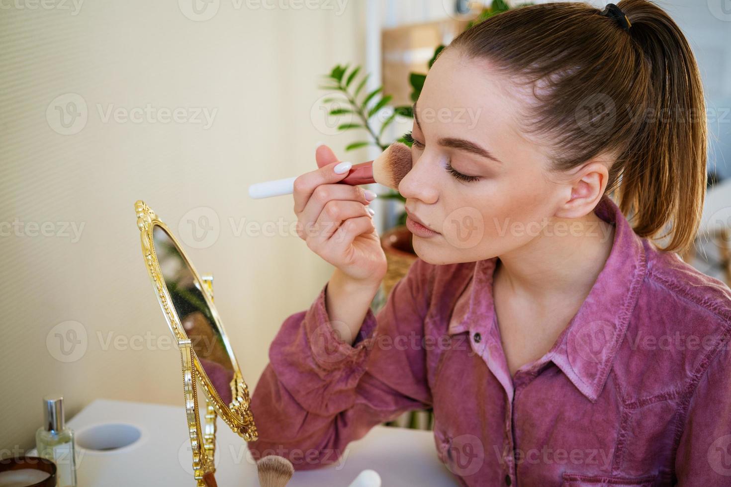 bela jovem faz blush no rosto usando pincel de maquiagem enquanto está sentado foto