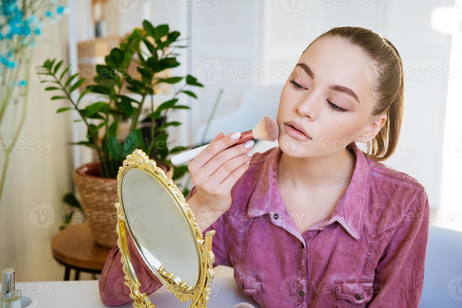 bela jovem faz blush no rosto usando pincel de maquiagem enquanto está sentado foto