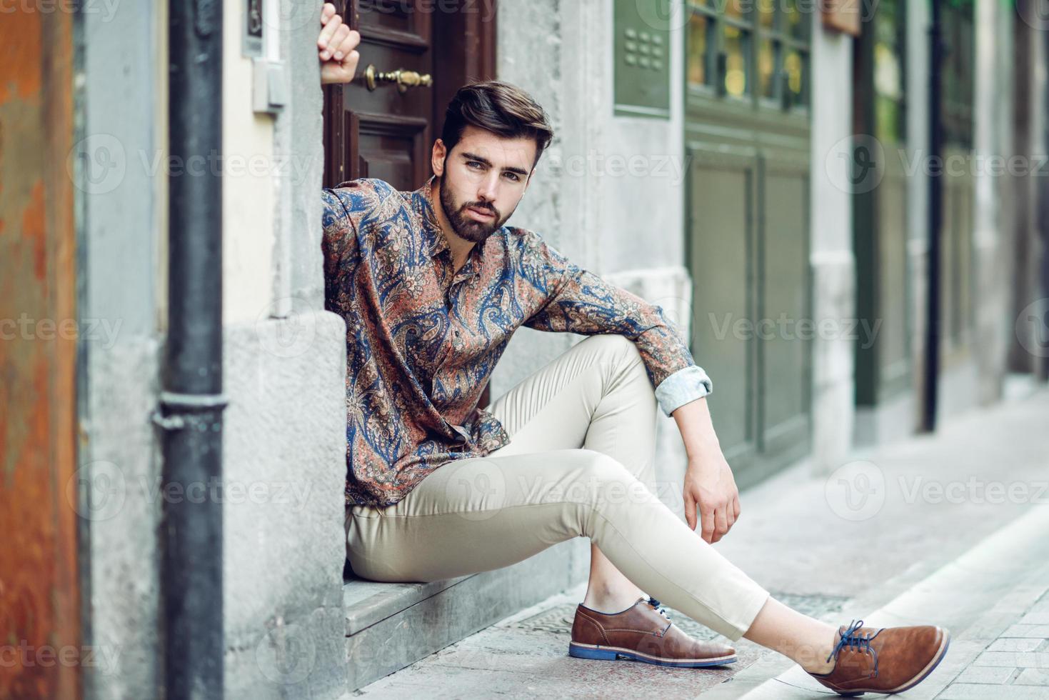 jovem barbudo, modelo de moda, sentado em um degrau urbano, vestindo roupas casuais. foto