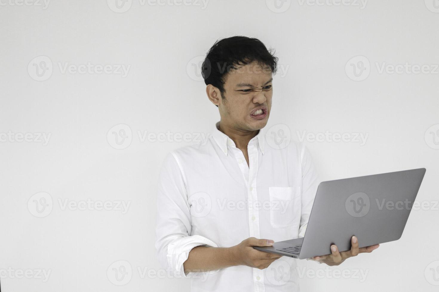 jovem asiático está com raiva e ódio com laptop. homem indonésia usar camisa branca isolado fundo cinza. foto