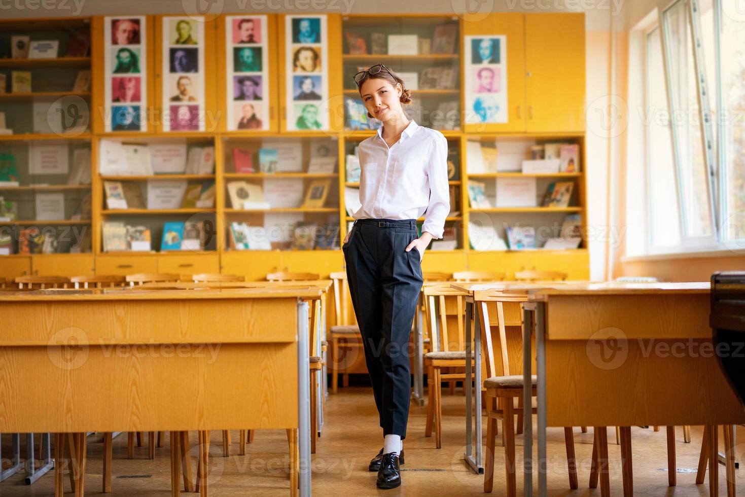 uma linda garota de camisa branca fica na sala de aula entre as mesas foto