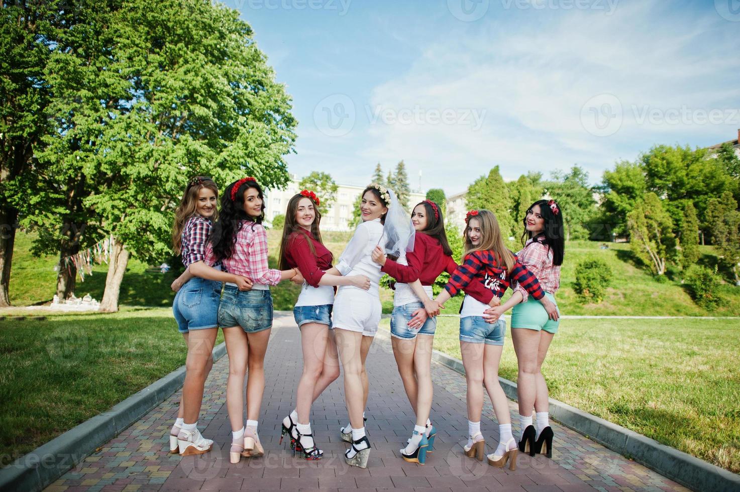 vista traseira de sete garotas felizes e sexy em shorts curtos de mãos dadas nas nádegas e posaram na estrada no parque na despedida de solteira foto