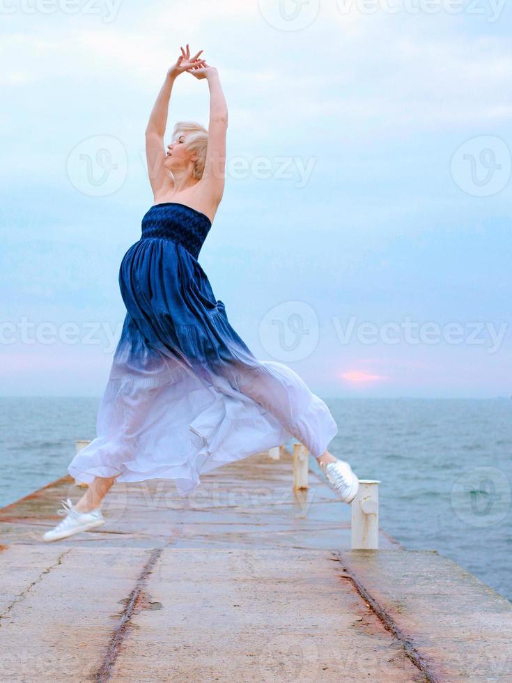 mulher grávida loira branca caucasiana com cabelo curto em vestido longo e tênis feliz por estar grávida e pulando à beira-mar ao nascer do sol foto