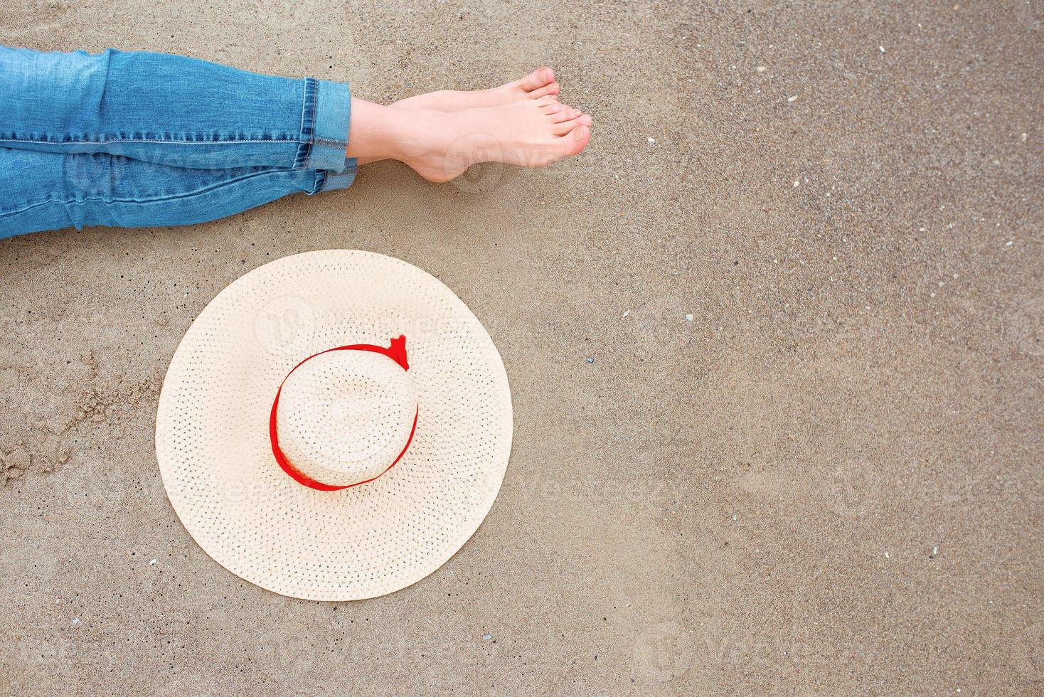 pernas e pés de mulher caucasiana em jeans sentado à beira-mar na costa com chapéu por perto no fundo de areia. foto abstrata de viagem