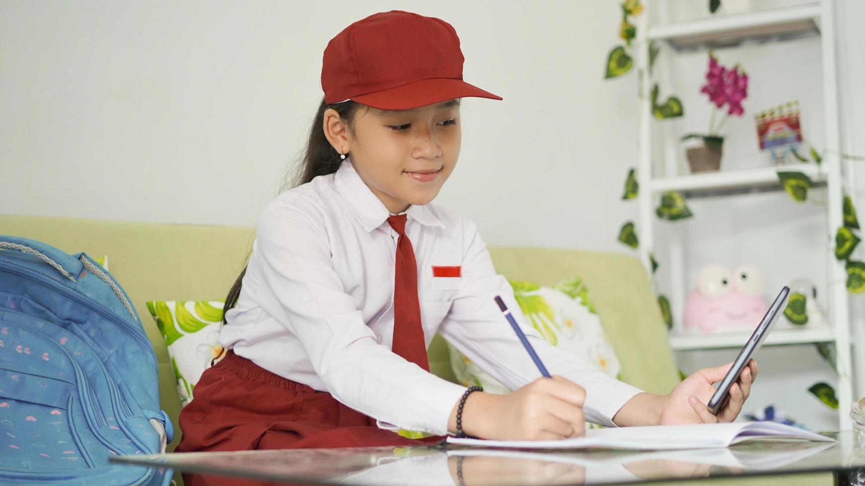 menina asiática da escola primária escrevendo ideias de seu smartphone em um livro em casa foto