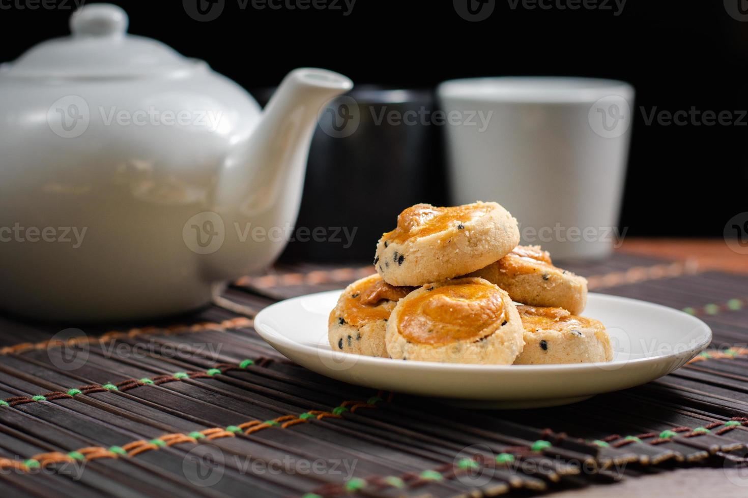 castanha de caju e biscoitos de gergelim preto no prato branco e mesa de madeira com bule de chá embaçado em fundo preto. conceito de chá da tarde. foto