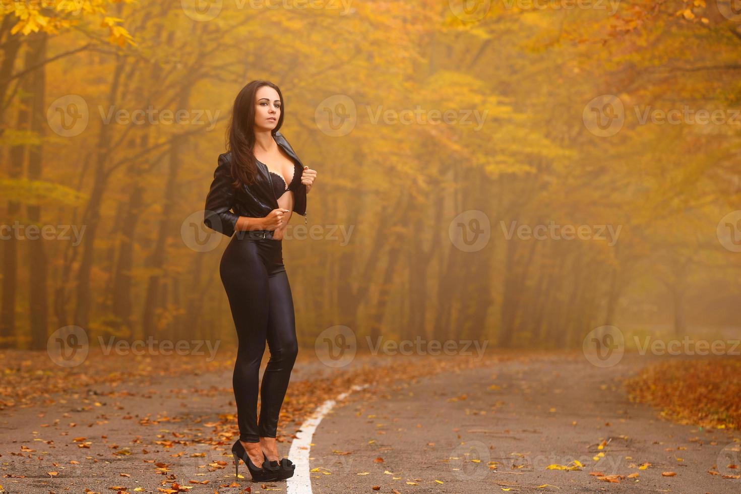 mulher jovem e bonita na floresta de outono na estrada posando de jaqueta preta e legging foto