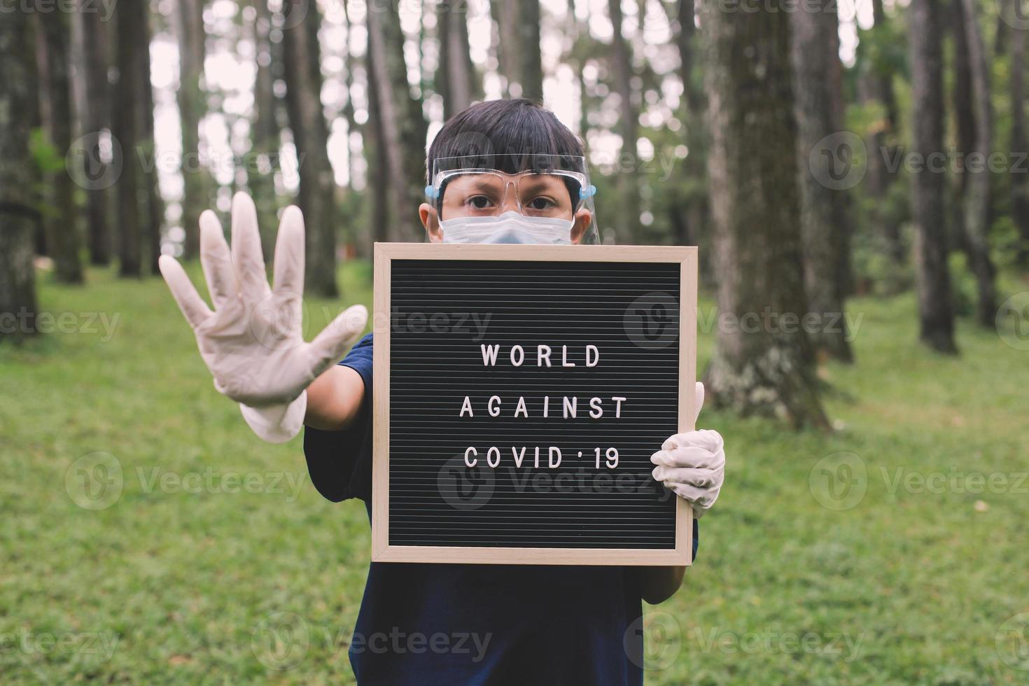 menino asiático usando máscaras faciais e luva de mão de borracha em pé com gesto de parada na mão enquanto segura citação no quadro de cartas diz mundo contra covid 19 foto