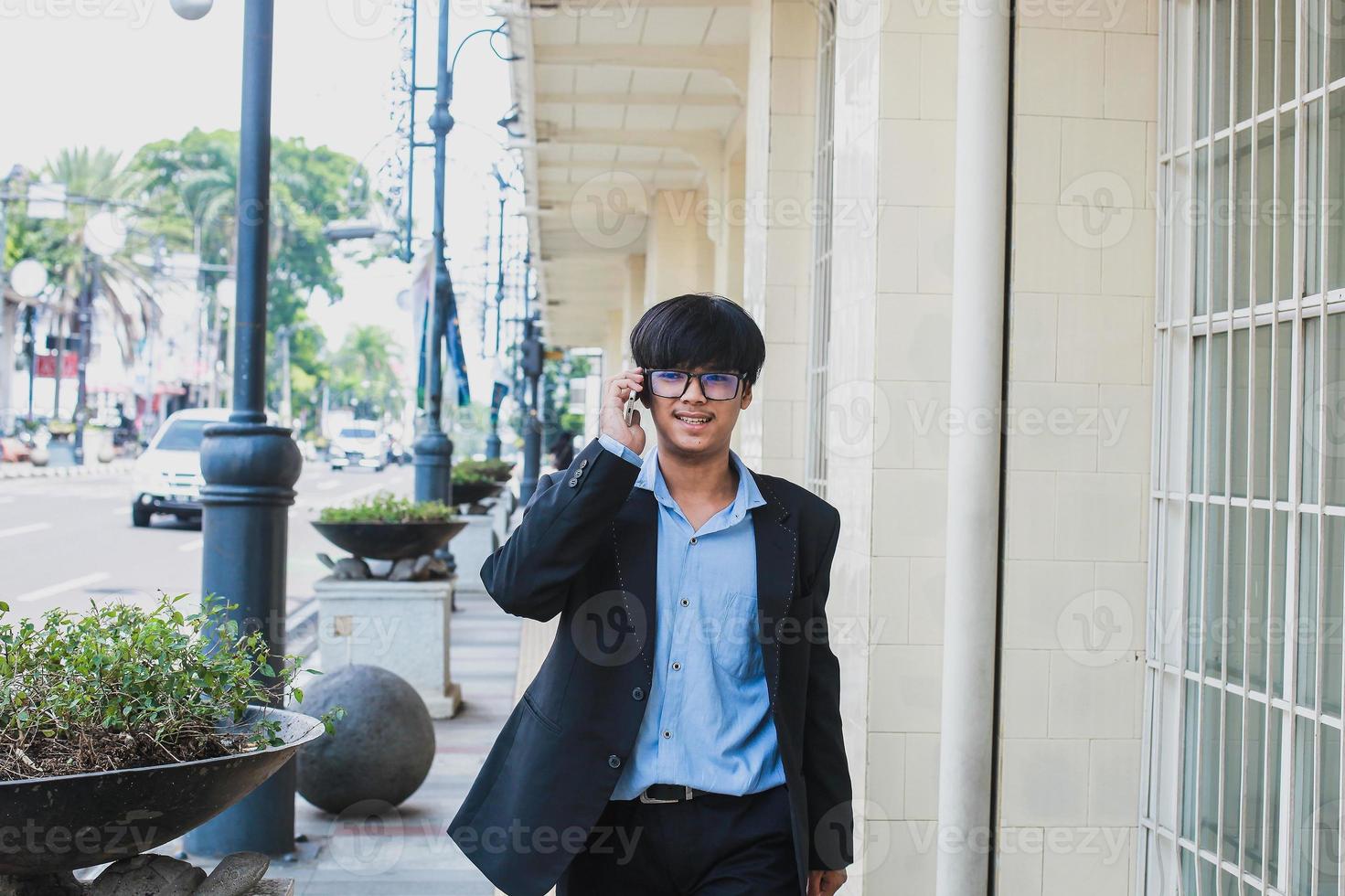 jovem asiático vestindo terno preto e óculos falar ao telefone enquanto sorrindo e andando foto