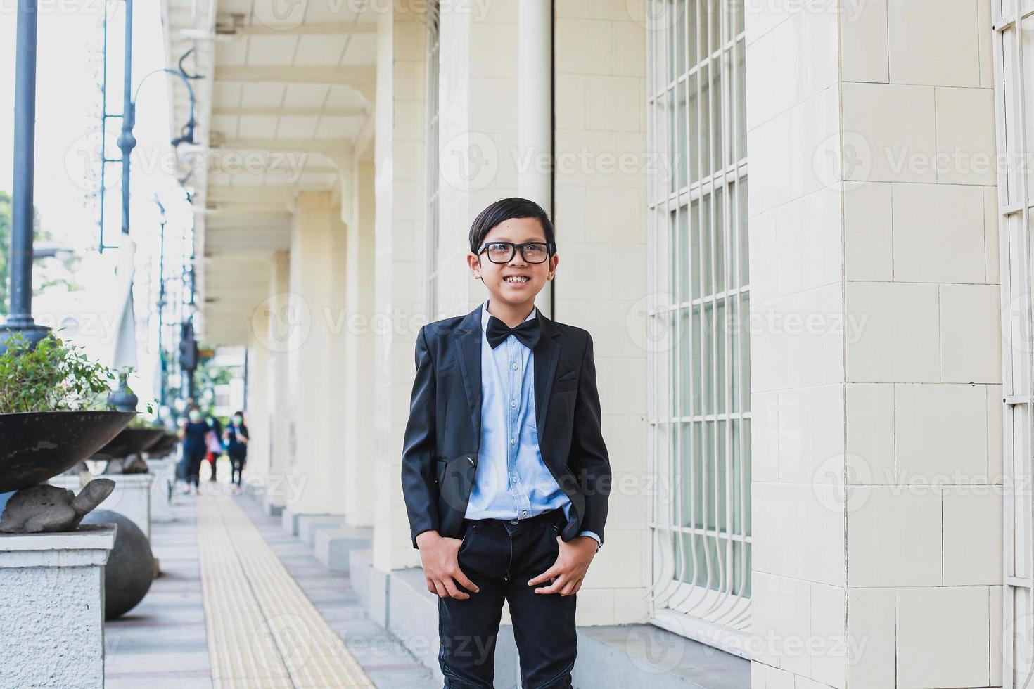 lindo menino asiático vestindo terno preto vintage e óculos posando e sorrindo para a câmera foto