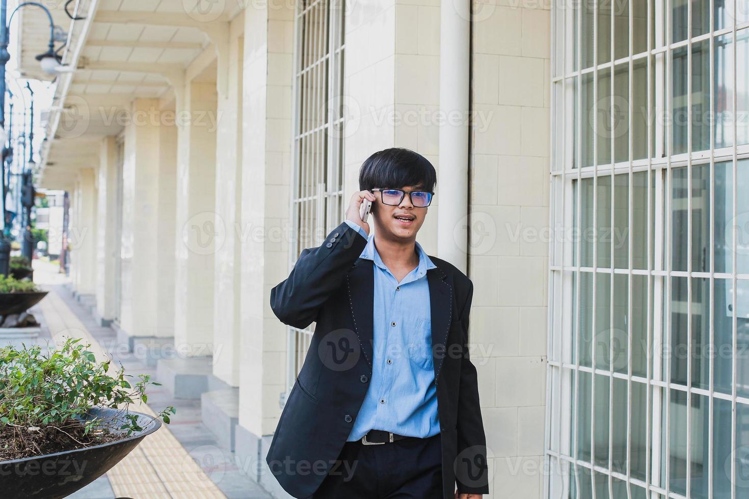 jovem asiático vestindo terno preto e óculos falar ao telefone enquanto sorrindo e andando foto