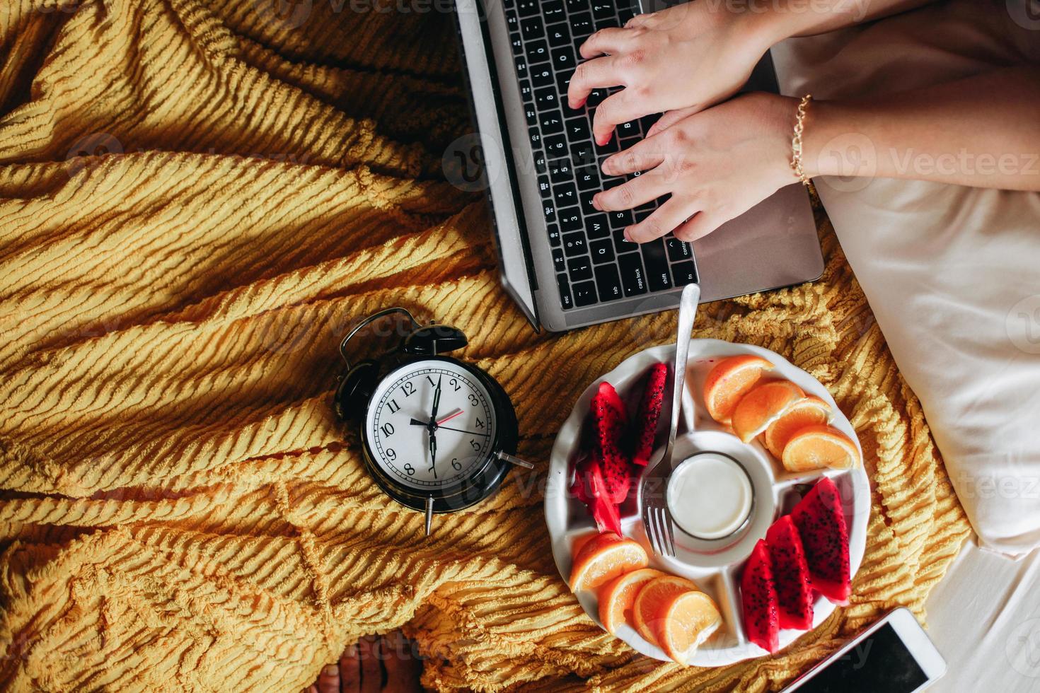 mão digitando no teclado do laptop com várias frutas no café da manhã e relógio mostrando 7 horas na cama foto