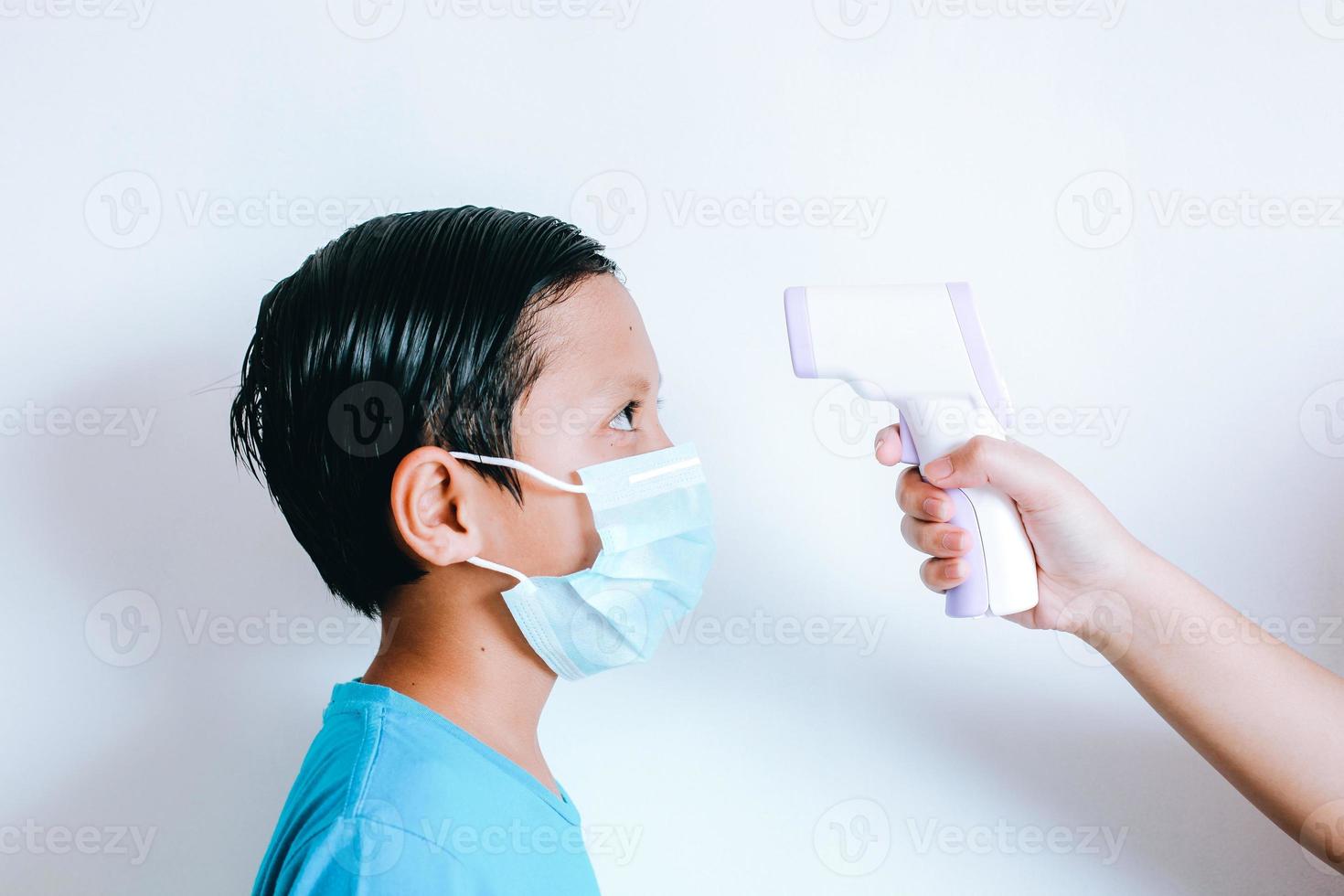 mão segurando o termômetro infravermelho medindo a temperatura do menino com máscara médica no rosto foto