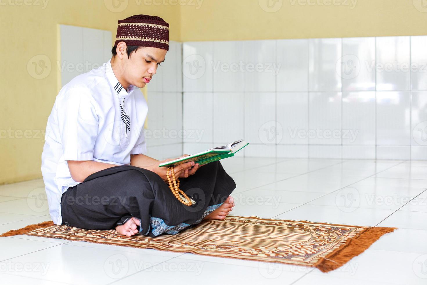 jovem muçulmano asiático segurando contas de oração e lendo o livro sagrado al-quran no tapete de oração foto