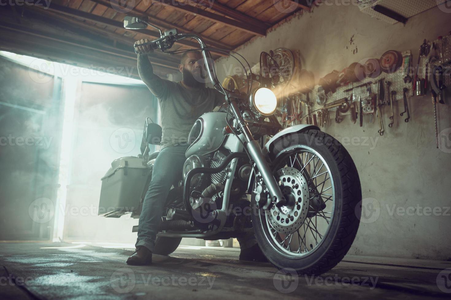 homem brutal bonito com barba sentado em uma motocicleta em sua garagem foto