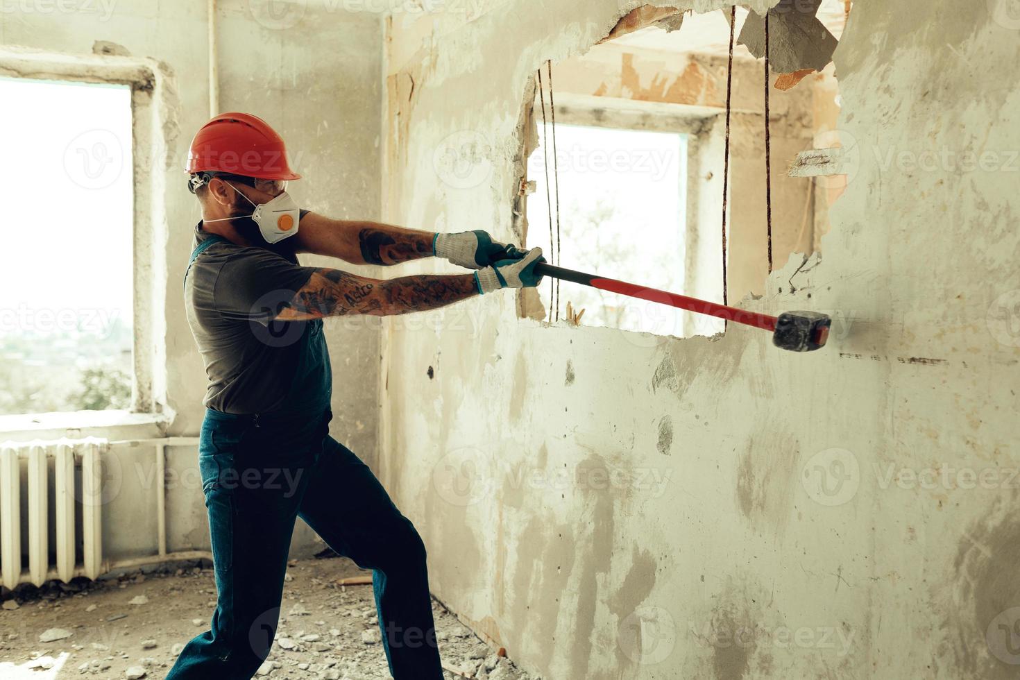 construtor com um martelo nas mãos quebra a parede de cimento o construtor está vestido com um traje de proteção e capacete foto