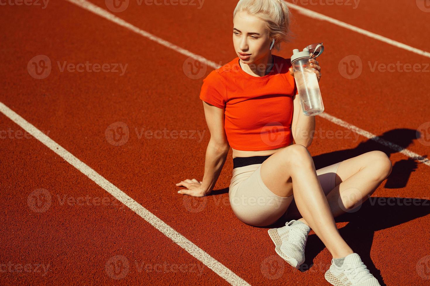 jovem loira bonita senta-se em uma pista de corrida com uma garrafa de água nas mãos foto