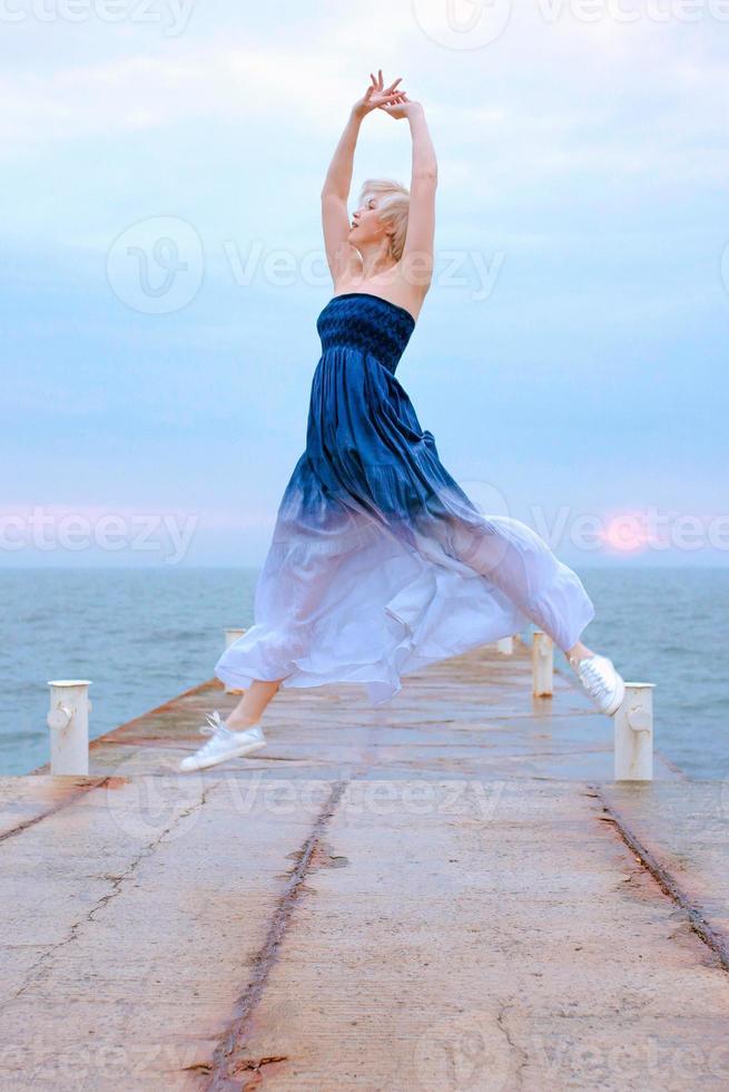 mulher loira branca caucasiana com cabelo curto em vestido longo e tênis feliz por estar grávida e pulando à beira-mar ao nascer do sol foto