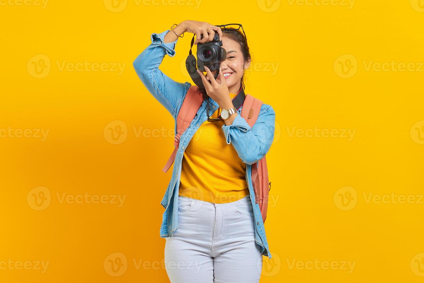 retrato de uma jovem asiática alegre com mochila tirando foto na câmera profissional isolada em fundo amarelo