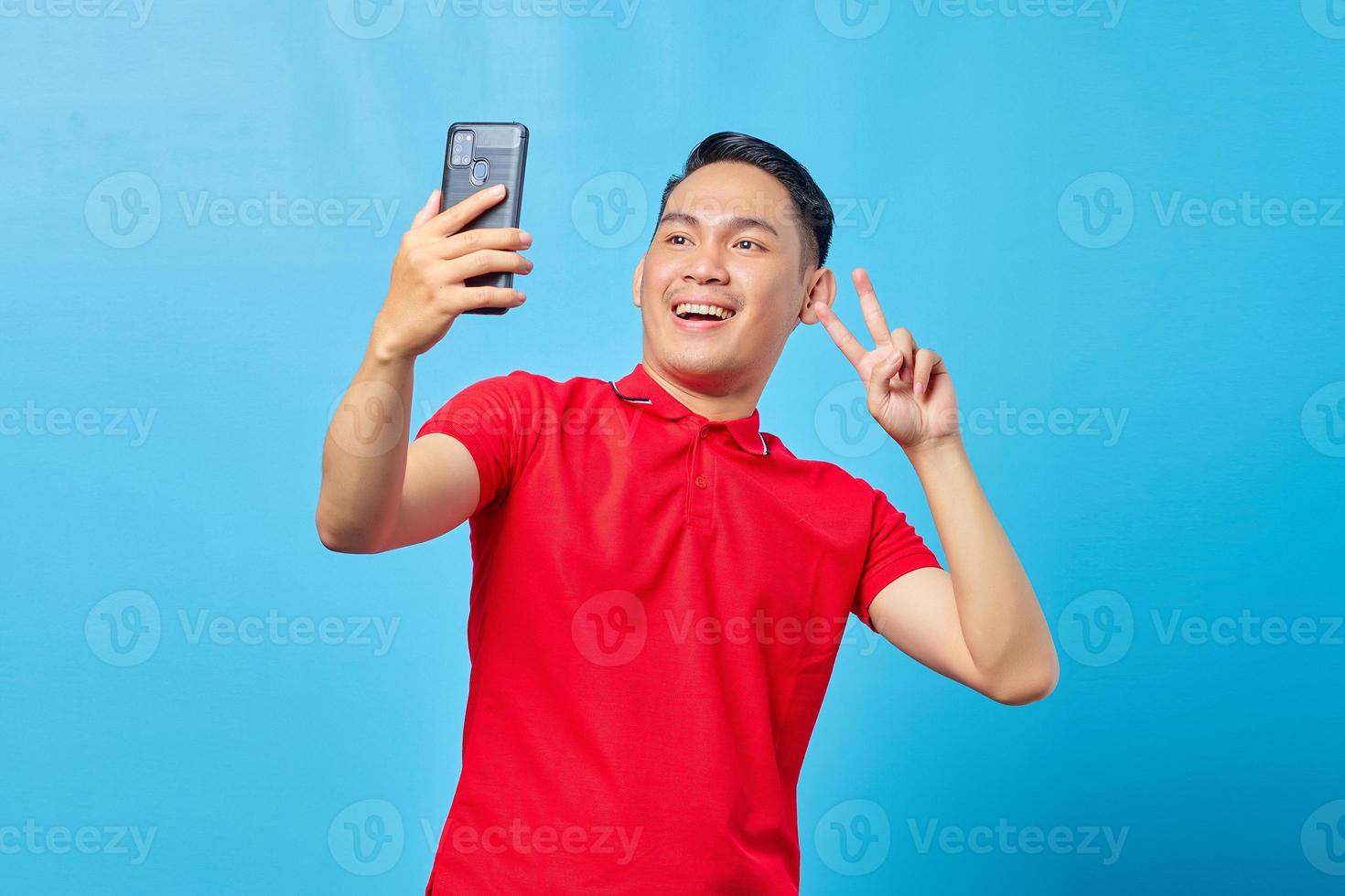 retrato de animado alegre jovem asiático usando telefone celular tirar selfie sobre fundo azul foto
