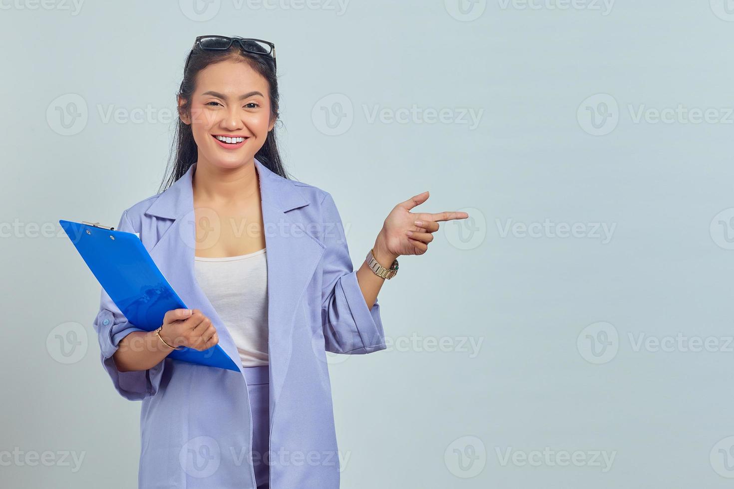 retrato de uma jovem asiática sorridente segurando a pasta de documentos e apontando o dedo para o espaço de cópia isolado no fundo roxo foto