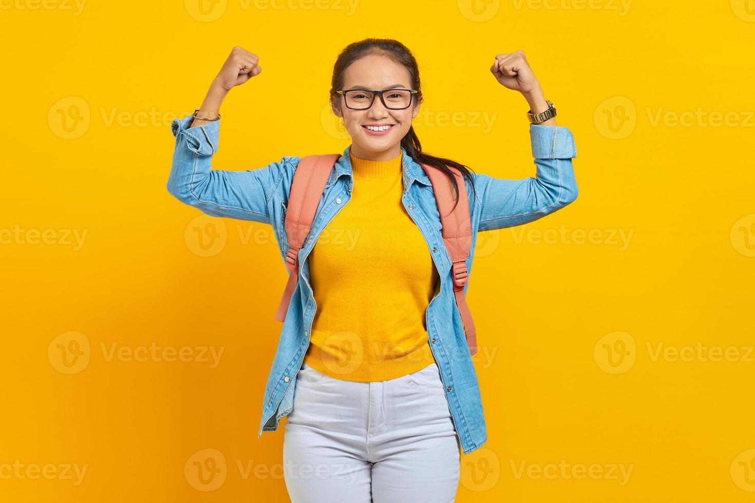 alegre jovem estudante asiática em roupa jeans com mochila levanta os braços e mostra o bíceps isolado em fundo amarelo. educação no conceito de universidade foto