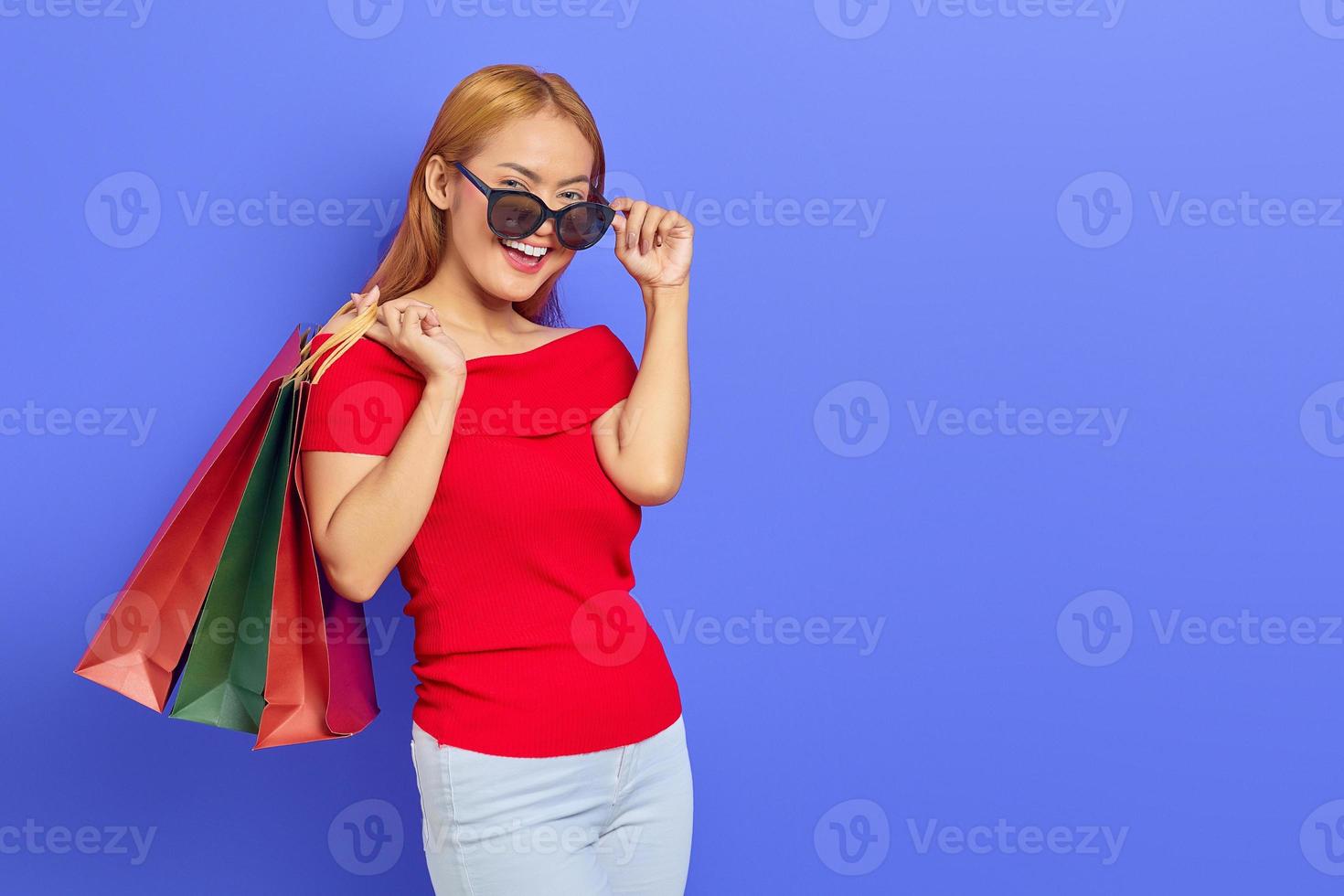 alegre linda mulher asiática de vestido vermelho e óculos segurando sacolas de compras isoladas sobre fundo roxo foto